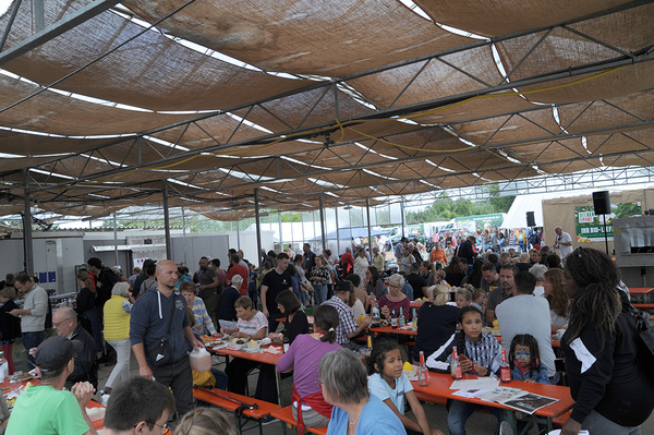 Eine große Gruppe von Menschen sitzt an Biertischen unter einer überdachten Fläche und isst. 