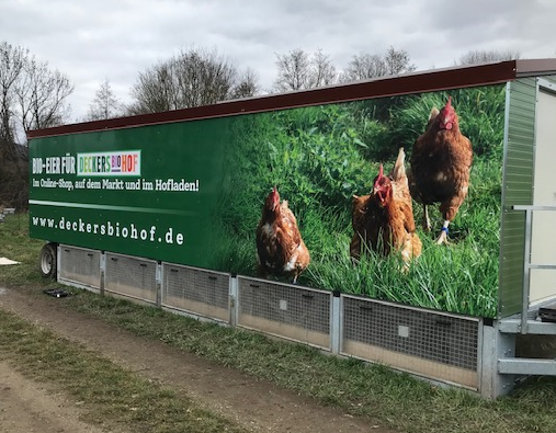 Ein großes, mobiles Hühnerstallgebäude (Hühnermobil) auf einem Feld. Auf der Seite des Stalls ist eine Plane mit einem großflächigen, fotorealistischen Bild von Hühnern in einer grünen Wiese zu sehen. Die Plane trägt das Logo "Deckers Biohof" und die Website-Adresse "deckersbiohof.de".