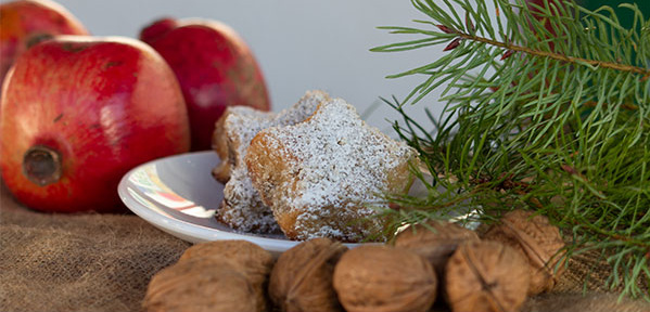  Eine festliche Nahaufnahme von zwei mit Puderzucker bestreuten, sternförmigen Plätzchen, die auf einem kleinen weißen Teller liegen. Im Hintergrund sind zwei rote Granatäpfel unscharf zu sehen. Rechts im Vordergrund liegen Walnüsse neben einem grünen Tannen- oder Wacholderzweig.