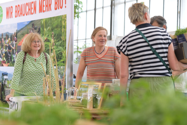 Drei Frauen stehen lachend an einem Verkaufsstand mit Pflanzen. Die Frau in der Mitte hält eine kleine Pflanze in der Hand. Ein Schild im Hintergrund ist teilweise sichtbar.